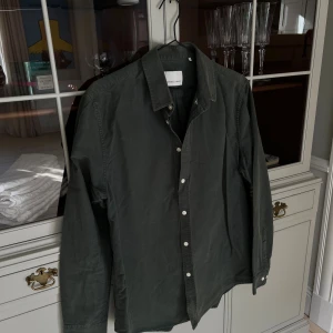 Mörkgrön skjorta från Samsøe - Stilren mörkgrön skjorta från Another Aspect i bomull. Klassisk krage, vita knappar framtill och långa ärmar. Perfekt för dig som gillar en enkel men snygg look. Skjortan har en rak passform och känns riktigt skön mot huden.
