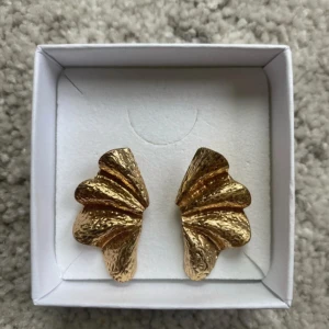 Guldiga vågformade örhängen - Unika örhängen i guldig färg med vågig, skulptural form och hamrad yta. De är iögonfallande och har en cool, konstnärlig vibe som verkligen sticker ut. Perfekta för dig som gillar statement-smycken och vill addera lite edge till din stil.