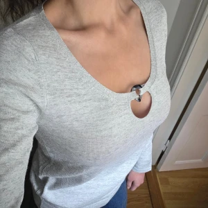 Grå långärmad tröja från Uterqüe - Grå långärmad tröja från Uterqüe i mjuk bomullsblandning. Tröjan har en rund halsringning med dekorativ ringdetalj framtill. Enkel och stilren modell som passar perfekt till jeans eller kjol.