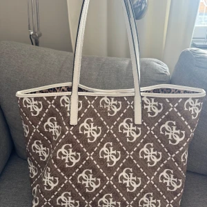 Brun shoppingväska från Guess med leopardfoder - Stor shoppingväska från Guess i brunt och vitt med logomönster över hela utsidan. Väskan har vita handtag och detaljer samt ett coolt leopardmönstrat foder på insidan. Perfekt för dig som vill ha plats för allt och samtidigt sticka ut med snygg design.