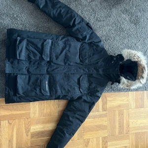 Svart dunjacka från Canada Goose - Säljer en svart dunjacka från Canada Goose med stor huva och snygg päls ( DSVDV). Jackan har flera stora fickor framtill och klassisk expedition-look. Perfekt för kalla dagar och riktigt stilren med sin mörka färg och rejäla passform.