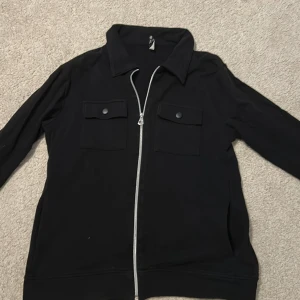 Svart overshirt med dragkedja - Svart overshirt med silverfärgad dragkedja framtill och två bröstfickor med lock och knapp. Jackan har klassisk krage och långa ärmar. Perfekt för lager-på-lager och enkel att styla till olika outfits. Storlek 176