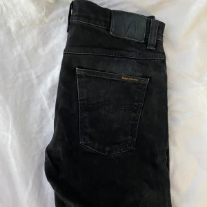 Svarta jeans från Nudie Jeans - Svarta jeans från Nudie Jeans med klassisk femficksdesign och diskret logga på bakfickan. Jeansen har raka ben och normal passform, tillverkade i slitstarkt bomullstyg. Perfekta för dig som gillar en stilren och enkel look.
