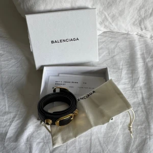 Balenciaga svart läderarmband med gulddetaljer - Svart armband från Balenciaga i äkta läder med chunky guldfärgade metalldetaljer och spänne. Armbandet levereras med original ask och dustbag samt kvitto. Perfekt accessoar för att lyfta din outfit.