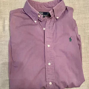 Lila skjorta från Polo Ralph Lauren - Snygg lila skjorta från Polo Ralph Lauren med klassisk button-down krage och grönt broderat logotyp på bröstet. Skjortan har långa ärmar, vita knappar framtill och är tillverkad i mjuk bomull.  Skjortan är oversized, snarare Large än Medium. Se mått på bild. Obs, bilderna med mått visar tvillingskjorta i annan färg. 