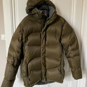 Olivgrön pufferjacka från Salomon - Fet olivgrön pufferjacka från Salomon med huva och stora, quiltade partier. Jackan har dragkedja framtill, två fickor med dragkedja och coolt tryck på insidan. Perfekt för kalla dagar när du vill hålla stilen och värmen.