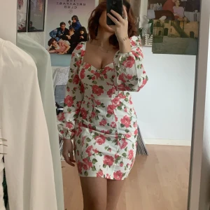 Vit blommig kortklänning med puffärm - Supersöt vit kortklänning med rosa blommor och gröna blad. Klänningen har puffiga långärmar, markerad byst och figurnära passform. Materialet känns som bomull och den har en dragkedja i ryggen.