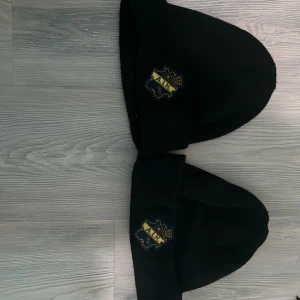 Svart AIK mössa med broderad logga - Svart mössa med AIK:s klassiska broderade klubbmärke framtill. Perfekt för dig som vill visa ditt stöd för laget och hålla dig varm. Enkel och stilren design med uppvikt kant. Passar både på läktaren och i vardagen.