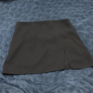 Svart kortkjol från Zara med slits - Stilren svart kortkjol från Zara i storlek L. Kjolen har en enkel design med en liten slits framtill och dragkedja i sidan. Perfekt för dig som gillar minimalistisk stil och vill ha en klassisk kjol i garderoben.