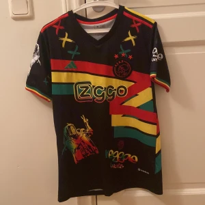 Ajax x Bob Marley (Limited Edition) - Unik Ajax fotbollströja från Adidas i samarbete med Bob Marley. Svart bas med färgglada ränder i rött, gult och grönt, samt tryck av Ziggo, Ajax-logga och reggae-inspirerade motiv. Korta ärmar med matchande färgdetaljer. Tillverkad i lätt och ventilerande material.