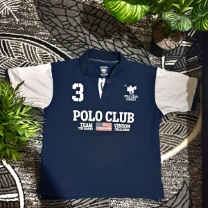 Mörkblå pikétröja Polo Club Vinson - Snygg mörkblå pikétröja från Vinson Polo Club med vita detaljer och tryck framtill. Tröjan har klassisk krage, korta ärmar i ljusgrått och broderad logga på bröstet. Perfekt för en sportig och stilren look.