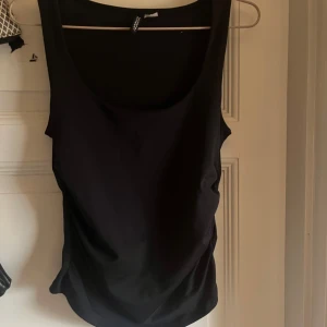 Svart linnetopp från H&M Divided - Svart ärmlös linnetopp från H&M Divided i storlek S. Toppen har en rundad urringning och snygga rynkade detaljer längs sidorna som ger en cool twist. Perfekt att styla med jeans eller kjol för en enkel men trendig look.