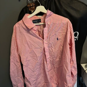 Rosa och vit randig skjorta Ralph Lauren - Klassisk rosa och vit randig skjorta från Ralph Lauren i bomull. Skjortan har lång ärm, knappar framtill och den ikoniska blå loggan broderad på bröstet. Classic fit-modell som passar perfekt till jeans eller chinos.