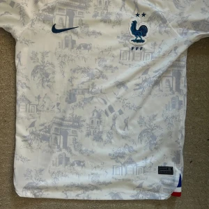 Frankrike Nike fotbollströja mönstrad - Säljer en officiell Frankrike fotbollströja från Nike i vitt med ljusgrått mönster av franska landmärken och motiv. Broderad tupp-logga och två stjärnor på bröstet, blå Nike-swoosh och trikolor-detalj i nederkant. Tillverkad i lätt och ventilerande material.
