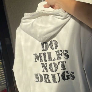 Vit hoodie med tryck ”do milfs not drugs” - Vit hoodie från Chicyea med svart statement-tryck på ryggen: 'DO MILFS NOT DRUGS'. Klassisk känguruficka framtill, huva med snörning och ribbade muddar. 
