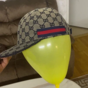 Gucci keps med monogram och röd-blå rand - Säljer en snygg Gucci keps i beige med klassiskt GG-monogram i mörkblått. Kepsen har en röd och blå rand på sidan och är tillverkad i bomullsmix. Perfekt accessoar för dig som vill sticka ut med lyxig streetstyle.