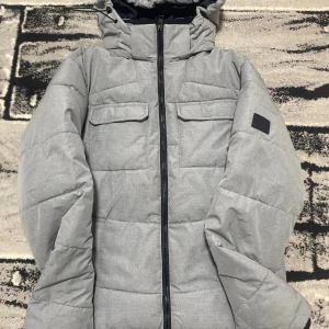 Grå pufferjacka Jack & Jones  - Snygg grå pufferjacka från Jack & Jones i storlek XS. Jackan har huva, två bröstfickor med lock och hel dragkedja framtill. Materialet är mjukt och quiltat, perfekt för kyliga dagar. Stilsäker och enkel att matcha med det mesta.