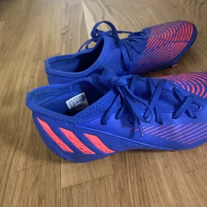 Adidas Predator blå fotbollsskor - Säljer ett par Adidas Predator fotbollsskor i blått med neonrosa detaljer och klassiska ränder på sidan. Skorna har snörning och är tillverkade i syntetmaterial för bästa grepp och känsla på planen. Perfekta för dig som vill sticka ut på matchen!