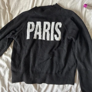Svart stickad tröja med PARIS-tryck - Svart stickad tröja med stor vit text 'PARIS' framtill. Tröjan har rund halsringning, ribbade muddar och lång ärm. Perfekt för dig som gillar streetstyle och vill ha en enkel men snygg statementtröja.