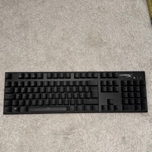 HyperX mekaniskt tangentbord - HyperX mekaniskt tangentbord i svart, fullstort med numerisk del. Tangenter och yta ser ut att vara i mycket gott skick utan synligt slitage. Perfekt för gaming eller arbete med snabb respons och hållbar konstruktion.