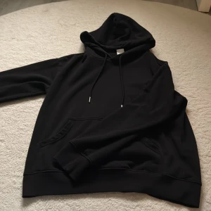 Svart hoodie i XS med huva - Svart hoodie i storlek XS med klassisk huva och dragsnören. Tröjan har en stor magficka och ribbade muddar vid ärmsluten. Perfekt för chill dagar eller när du vill ha en enkel och stilren look. 