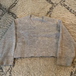 Grå stickad croppad tröja V-ringad - Grå stickad tröja med V-ringning och croppad passform. Tröjan har bred ribbad mudd vid ärmslut och nederkant, vilket ger en snygg och modern look. Perfekt för lager-på-lager under hösten och vintern.