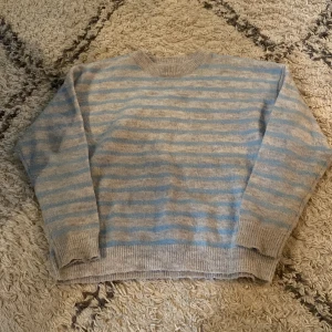 Randig stickad tröja i blått och beige - Mysig stickad tröja med ljusblå och beigea ränder. Tröjan har rund halsringning, ribbade muddar och lång ärm. Perfekt för lager-på-lager och chill dagar.