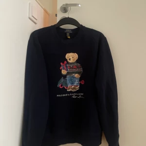 Mörkblå Polo Bear tröja från Ralph Lauren - Säljer en mörkblå sweatshirt från Ralph Lauren med ikoniska Polo Bear-trycket framtill. Björnen har stickad tröja, jeans och håller ett paket med röd rosett. Tröjan har rund hals och långa ärmar, perfekt för dig som vill ha en klassisk men lekfull stil. Storlek Xl barn men passar även storlek S