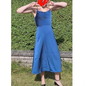 Blå långklänning med smala axelband - Snygg blå långklänning med smala axelband och lätt utställd passform. Klänningen har en enkel och stilren design som ger en elegant siluett. Materialet är mjukt och har ett lätt glansigt utseende, perfekt för varmare dagar. Behövs du lite på ena axelbandet. Över det är den som ny 