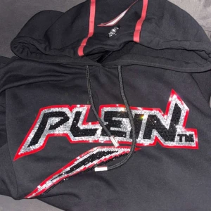 Svart hoodie från Philipp Plein med paljetter - Svart hoodie från Philipp Plein med stor logga framtill i silver- och svarta paljetter, röd kant och coola röda detaljer på huvan. Tröjan har snörning i halsen och är perfekt för dig som vill sticka ut med en streetstyle-look.