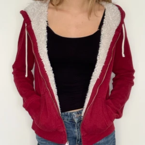 Röd teddyhoodie med huva från Hollister - Mysig röd teddyhoodie från Hollister med vit fluffig fuskpäls på insidan och huva. Jackan har dragkedja, snörning i huvan och två fickor framtill. 