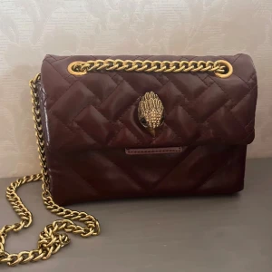 Quiltad vinröd axelväska Kurt Geiger - Snygg vinröd axelväska från Kurt Geiger med quiltad design och guldfärgad kedja. Väskan har ett coolt fågelhuvud som spänne och en metallbricka med logga. Tillverkad i skinn och har ett rymligt fack invändigt. Perfekt för dig som vill sticka ut med detaljer. Tryck ej på köp nu!!! 