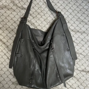 Grå axelväska från KappAhl - Stilren grå axelväska från KappAhl i skinnimitation med flera dragkedjefickor framtill och justerbar axelrem. Väskan har en rymlig insida med innerfack och snygga metalldetaljer. 👍🏼😊