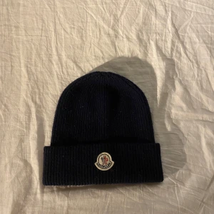 Mörkblå Moncler mössa i ull - Snygg mörkblå ribbstickad mössa från Moncler med klassisk logotyp framtill. Mössan är tillverkad i mjuk ull och har en uppvikt kant för extra stilpoäng. Perfekt accessoar för kyliga dagar och en clean look.