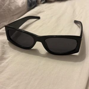 Svarta solglasögon från Saint Laurent - Snygga svarta solglasögon från Saint Laurent med rektangulära bågar och guldigt YSL-logo på sidan. Tillverkade i Italien med en stilren och chunky design som ger en cool vibe. Perfekta för dig som vill sticka ut med en lyxig accessoar.
