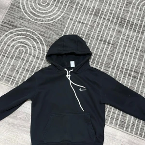 Svart Nike hoodie med vit snörning - Svart hoodie från Nike med vit snörning och klassisk swoosh-logga på bröstet. Tröjan har huva, känguruficka och långa ärmar. Perfekt för en chill och sportig look.