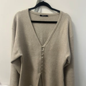 Beige kofta med knappar från Gina Tricot - Snygg beige kofta från Gina Tricot med djup v-ringning och dekorativa knappar framtill. Koftan har lång ärm och är i mjukt material som känns skönt mot huden. Perfekt att slänga på sig när det är lite kyligare ute. Detta är en väldigt populär tröja som ofta är slutsåld. Den är i storlek M och är använd ett fåtal gånger. 