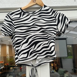 Zebramönstrad croppad topp med knyt - Säljer en kort zebramönstrad topp i svart och vitt med knyt i midjan och öppen rygg. Toppen har rund halsringning och korta ärmar. Perfekt för dig som vill sticka ut med ett djurmönster och cool detalj i ryggen.