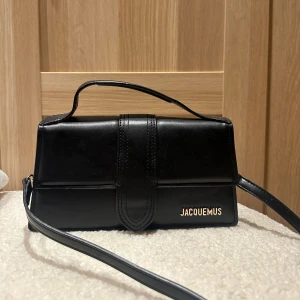 Svart axelväska från Jacquemus - Snygg svart axelväska från Jacquemus i stilren, rektangulär form. Väskan är tillverkad i slätt skinn och har ett elegant handtag samt axelrem. Guldiga Jacquemus-bokstäver framtill ger en lyxig touch. Perfekt för dig som gillar minimalistisk design.