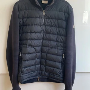Svart cardigan från Moncler - Snygg svart cardigan från Moncler med quiltad framsida och släta ärmar. Jackan har dragkedja framtill, två fickor med dragkedja och ribbad krage. Perfekt för kyliga dagar och ger en clean look.