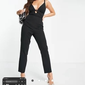 Svart jumpsuit med smala axelband - Säljer en svart jumpsuit från ASOS med smala axelband och djup v-ringning framtill. Byxorna är raka och har en croppad längd. Snygga banddetaljer över bysten ger en extra touch. Perfekt för dig som vill sticka ut med en stilren look. Helt oanvänd 
