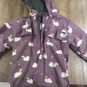 Lila regnset Petite Chérie Atelier 130 - Superfin lila regnjacka från Petite Chérie Atelier i storlek 130. Jackan har ett gulligt svanmönster med små rosa kronor, huva och dragkedja framtill. Insidan är fodrad med mjuk fleece för extra komfort. Perfekt för regniga dagar! Matchande regnbyxor med hängslen, också fodrade! 