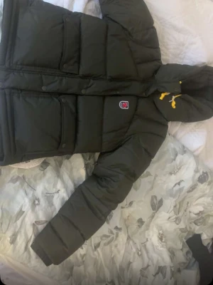 Svart pufferjacka Fjällräven Expedition - Säljer en svart pufferjacka från Fjällräven Expedition Series 1974 med huva och gula snören. Jackan har stora fickor med knappar och klassisk Fjällräven-logga på bröstet. Perfekt för kalla dagar. den är helt ny och har knappt andvänt den