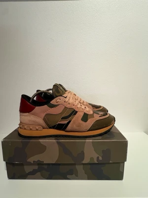 Valentino Rockrunner sneakers camouflage - Säljer ett par Valentino Rockrunner sneakers med snyggt kamouflagemönster i grönt, beige och brunt. Passar från 39-41  Skorna har detaljer i mocka och skinn, samt en röd häl med nitar. Platt sula och rund tå, med snörning framtill. Skicket är som på bild är i perfekt skick endast lite färg som försvunnit från framsidan av skon men knappts märks| medföljs dustbag box+ extra snören| fraktas den 2 januari 