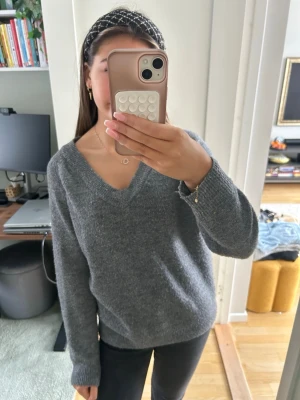 Grå stickad v-ringad tröja - Mysig grå stickad tröja med v-ringning och långa ärmar. Perfekt för kyligare dagar och enkel att matcha med jeans eller kjol. Tröjan har en klassisk passform och ribbade muddar vid ärmslut och nederkant.