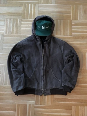 Vintage Carhartt Active Jacket J130 Washed Duck Canvas St M - Säljer en mörkgrå hoodiejacka från Carhartt Active Jacket  med dragkedja, snörning i huvan och två stora fickor framtill. Jackan har ribbade muddar och är tillverkad i slitstark bomull. Perfekt för dig som gillar streetwear och vill ha en chill look. OBS! Kepsen ingår inte!