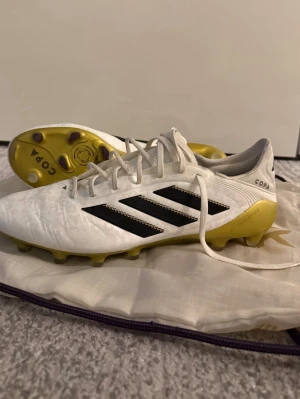 Adidas Copa vita fotbollsskor - Säljer ett par vita Adidas Copa fotbollsskor med svarta detaljer och gula dobbar. Skorna har snörning och är tillverkade i syntetmaterial med mönstrad ovandel. Perfekta för dig som vill sticka ut på planen med stilrena och sportiga skor.
