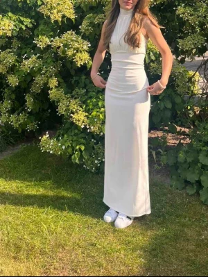 Vit långklänning med hög hals - Säljer en elegant vit långklänning med hög hals och ärmlös design. Klänningen har en figurnära passform och en snygg slits! Materialet känns mjukt och följsamt mot kroppen. Använd en gång ❤️