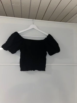Svart puffärm topp Gina Tricot M - Säljer en gullig svart topp från Gina Tricot i storlek M. Modellen har puffiga korta ärmar och smockad kropp som ger en tight och bekväm passform. Toppen är croppad och har en offshoulder-design. Perfekt till jeans eller kjol😊⭐️
