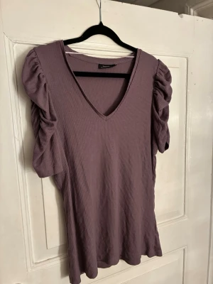Lila ribbad topp med puffärm KappAhl - Säljer en lila ribbad topp från KappAhl med v-ringning och korta puffärmar. Toppen har en loose passform och är tillverkad i mjukt, stretchigt material. Perfekt för dig som gillar voluminösa ärmar och en trendig look.